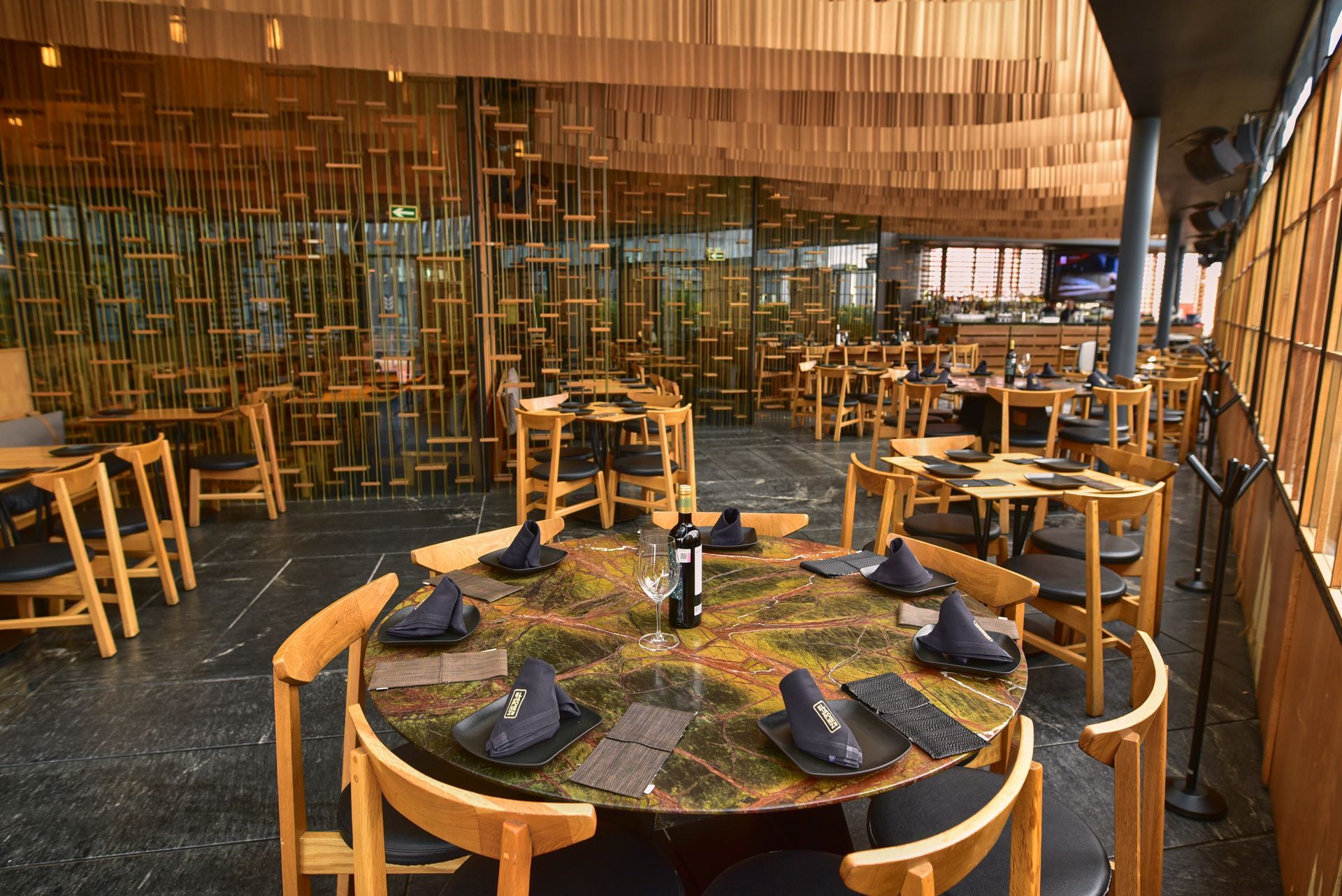 Terraza de restaurante al aire libre con mesa redonda, botellas y servilletas dobladas, rodeada de sillas de madera bajo una iluminación cálida.