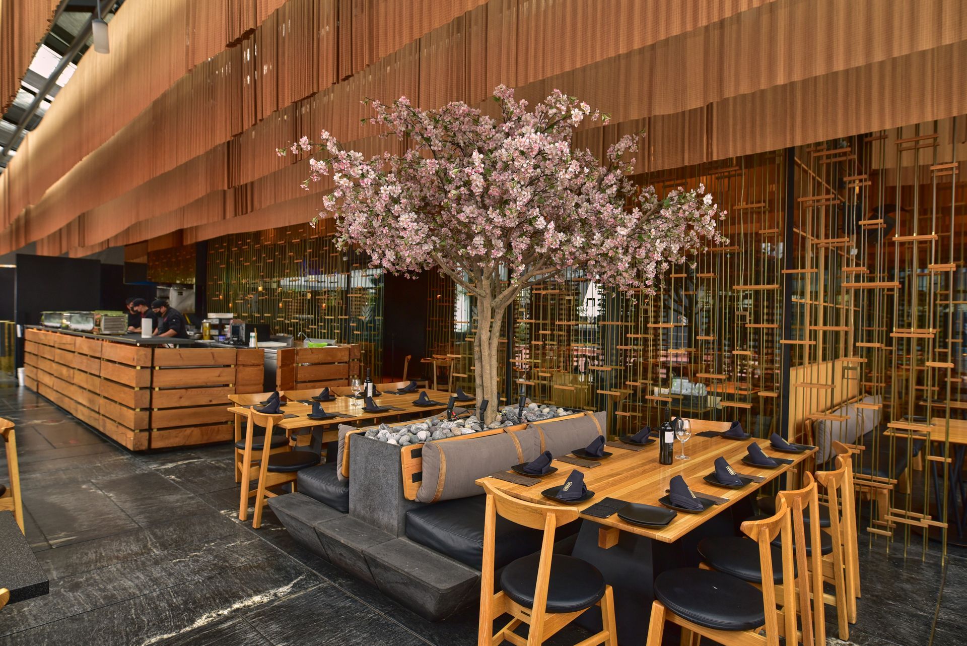 Patio de restaurante moderno con mesas de madera, sillas negras y un árbol de flores rosas bajo un techo curvo de cobre.
