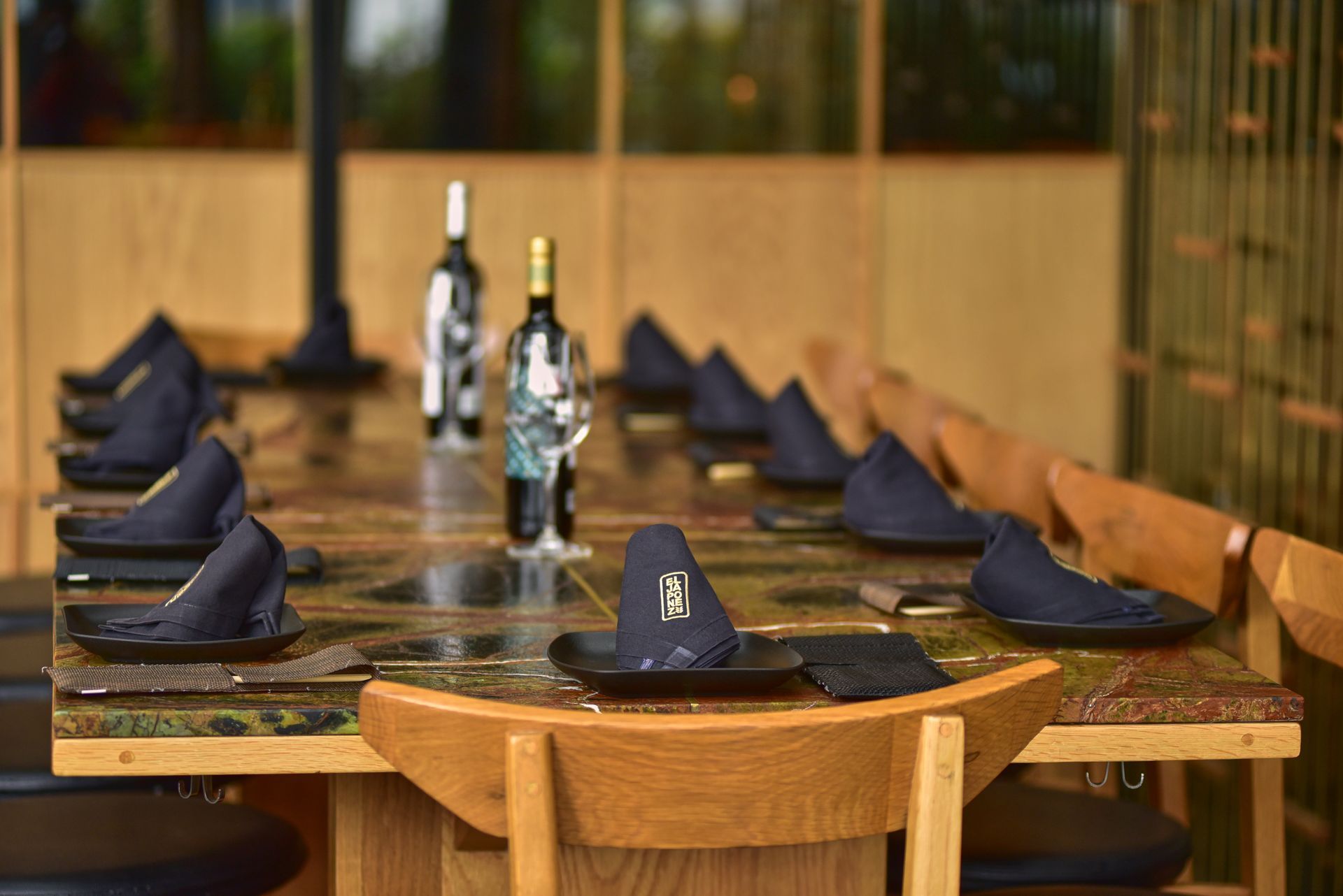 Prepara la mesa con servilletas dobladas, vasos y botellas de vino en un ambiente de restaurante de tonos cálidos.