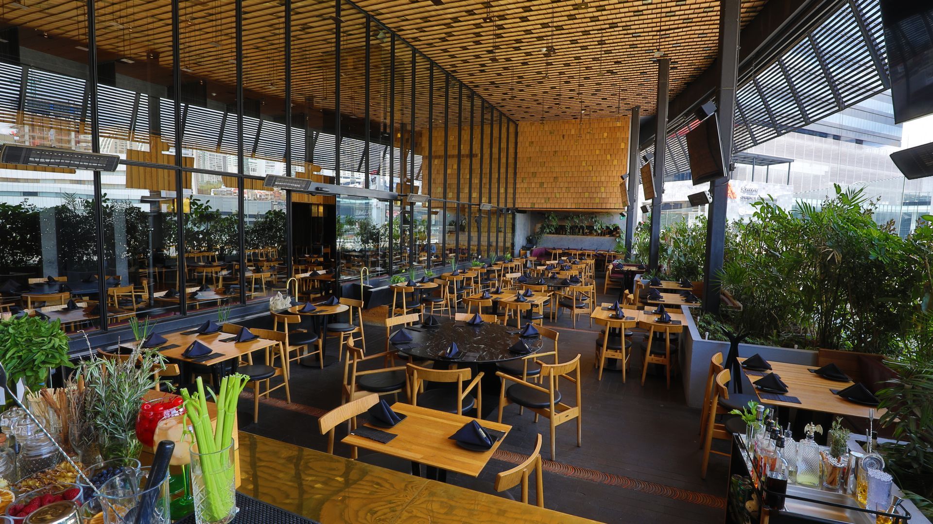 Restaurante al aire libre con techo de madera, mesas negras y amarillas y plantas en macetas.