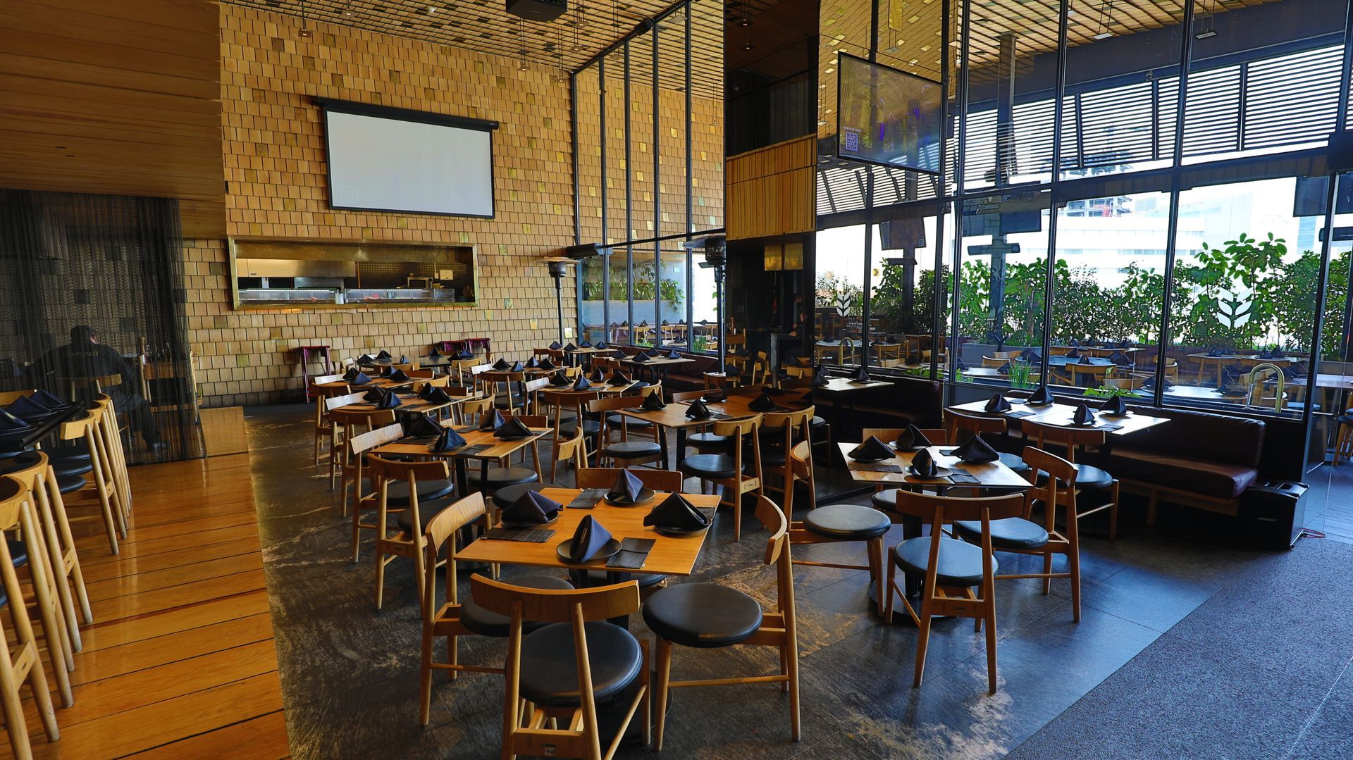 Interior de restaurante moderno con mesas y sillas de madera, grandes ventanales e iluminación cálida.