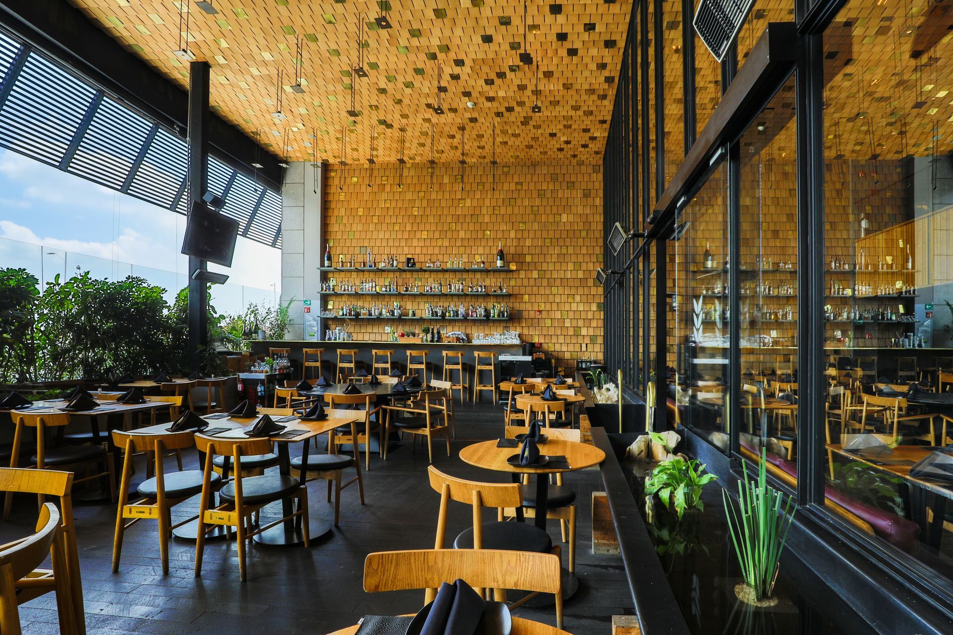 Interior de restaurante moderno con mesas de madera, sillas negras, paredes de cristal y un techo con un cálido estampado.