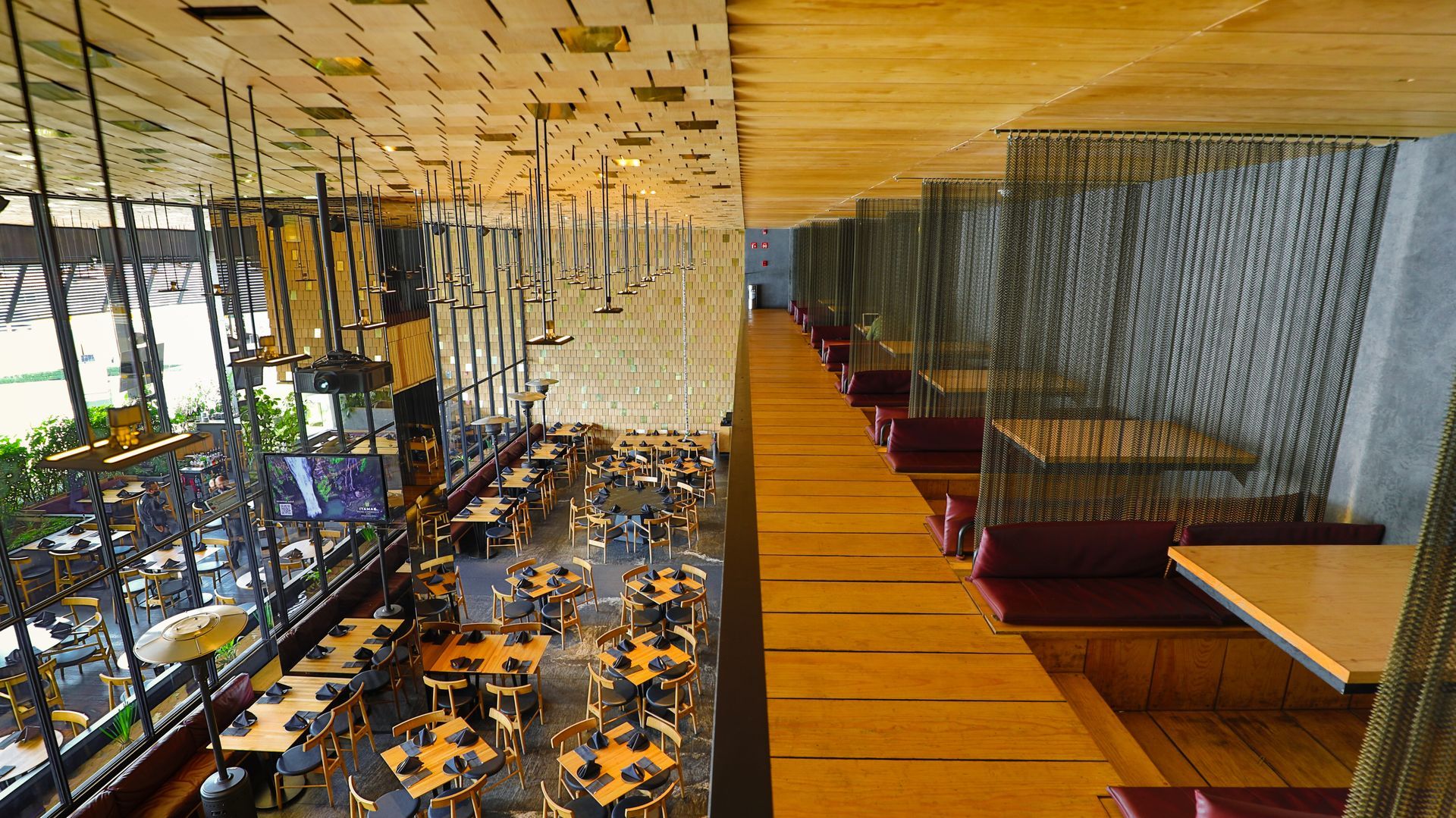 Interior de restaurante moderno con pasarela de madera, lámparas colgantes y mesas junto a grandes ventanales.