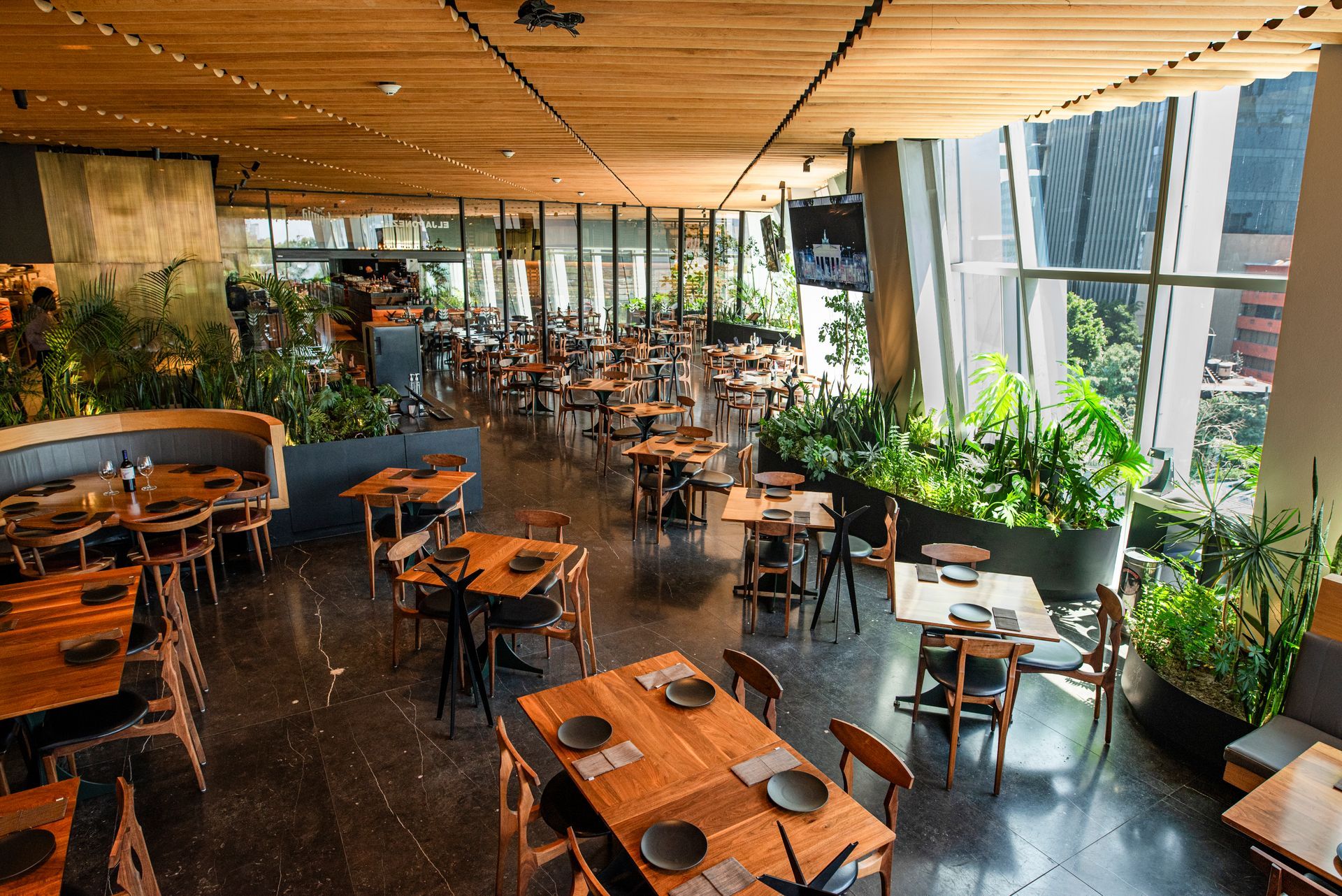 Interior de restaurante moderno con mesas y sillas de madera, y grandes ventanales con vistas a la vegetación.
