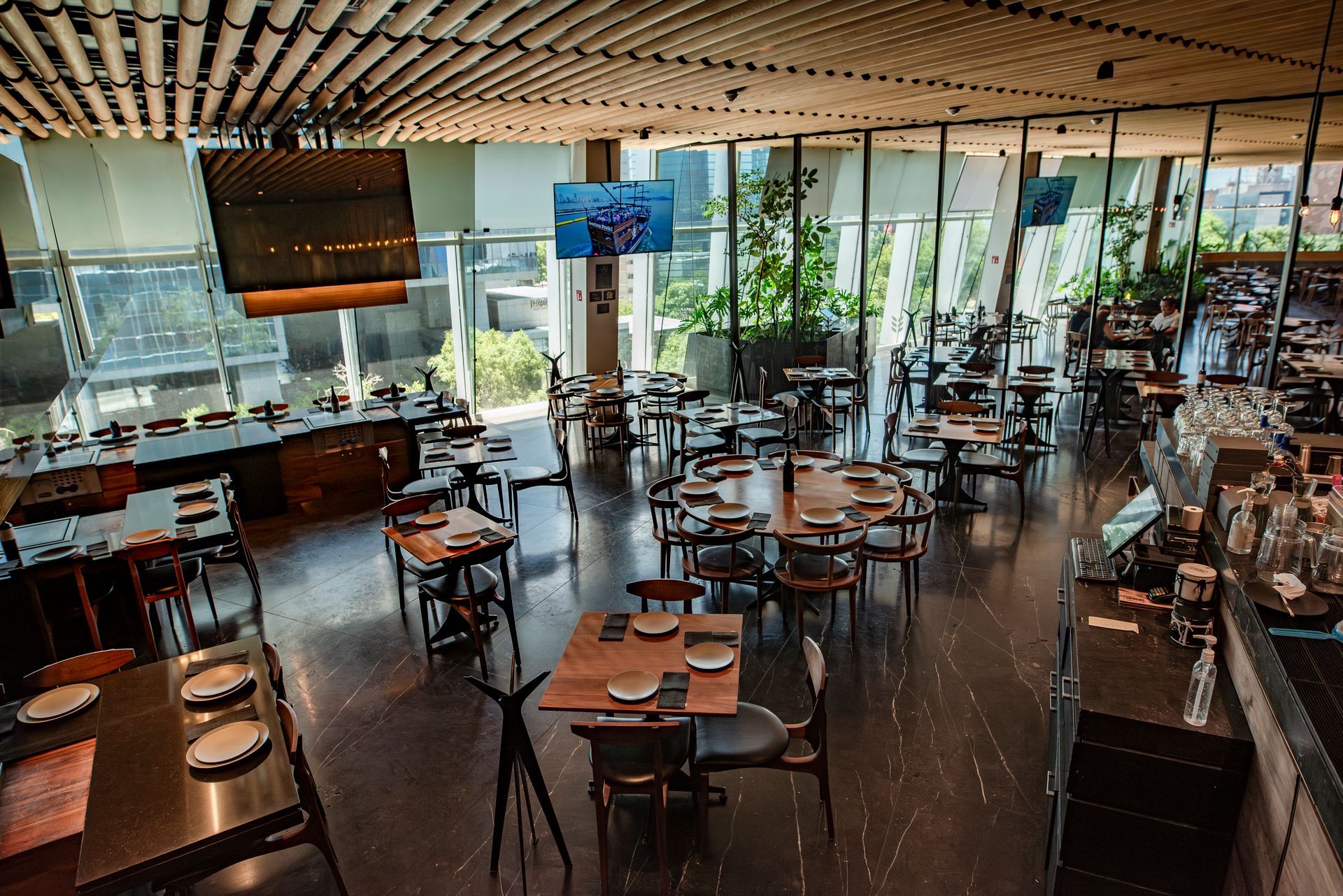 Cafetería moderna con mesas y sillas vacías, y grandes ventanales con vistas a zonas verdes.