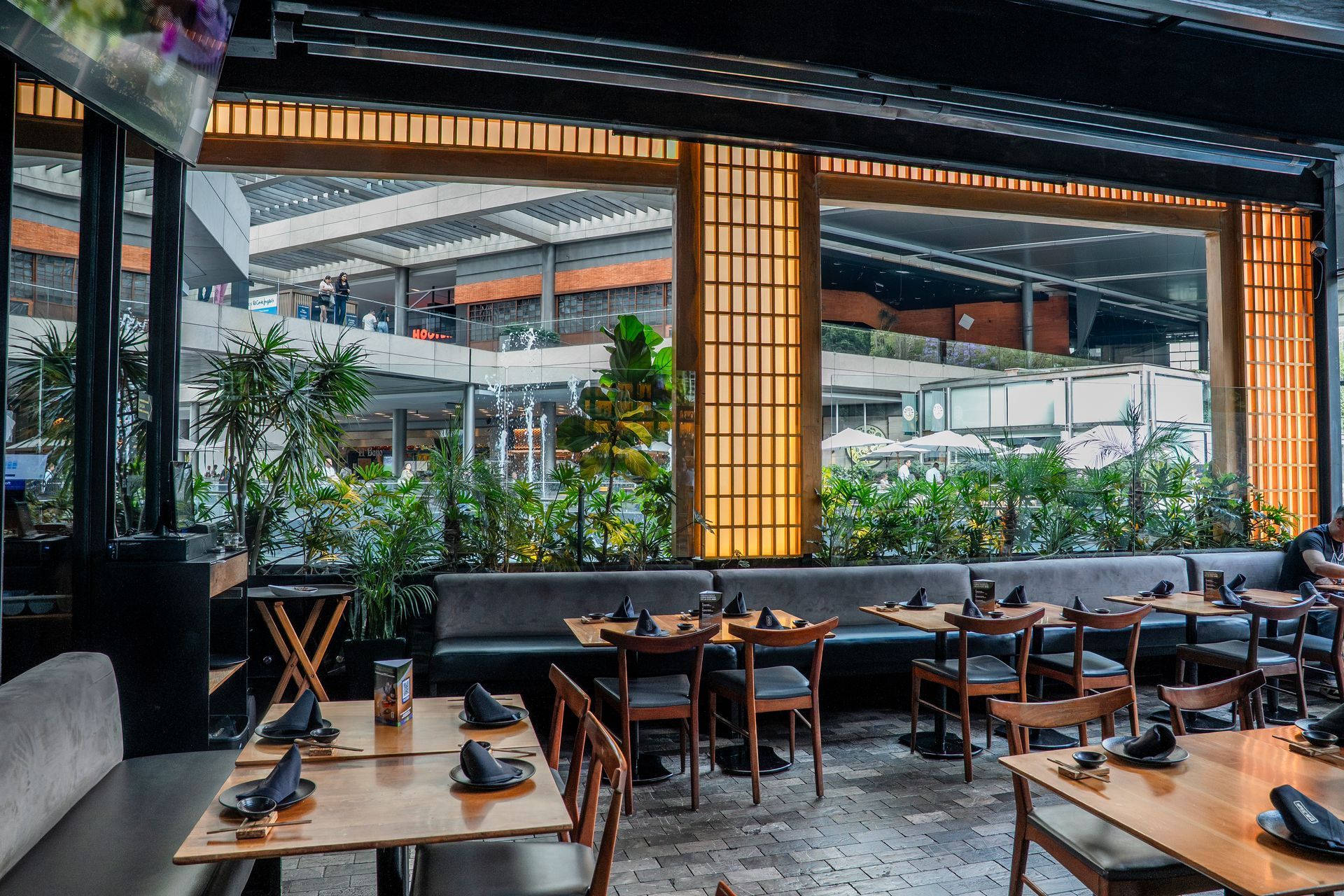 Elegante restaurante al aire libre con mesas y sillas de madera y exuberantes plantas bajo un techo de metal.