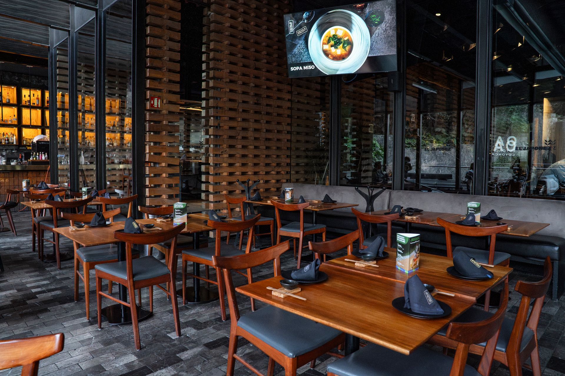 Comedor vacío de un restaurante moderno con mesas de madera, sillas oscuras y una pared llena de botellas de vino.