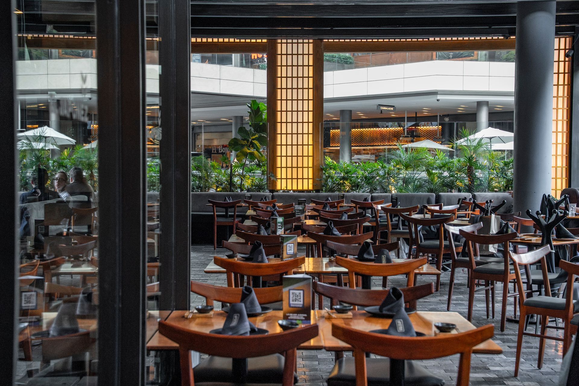 Patio de restaurante moderno con mesas y sillas de madera, faroles cálidos y exuberante vegetación bajo un techo cubierto.