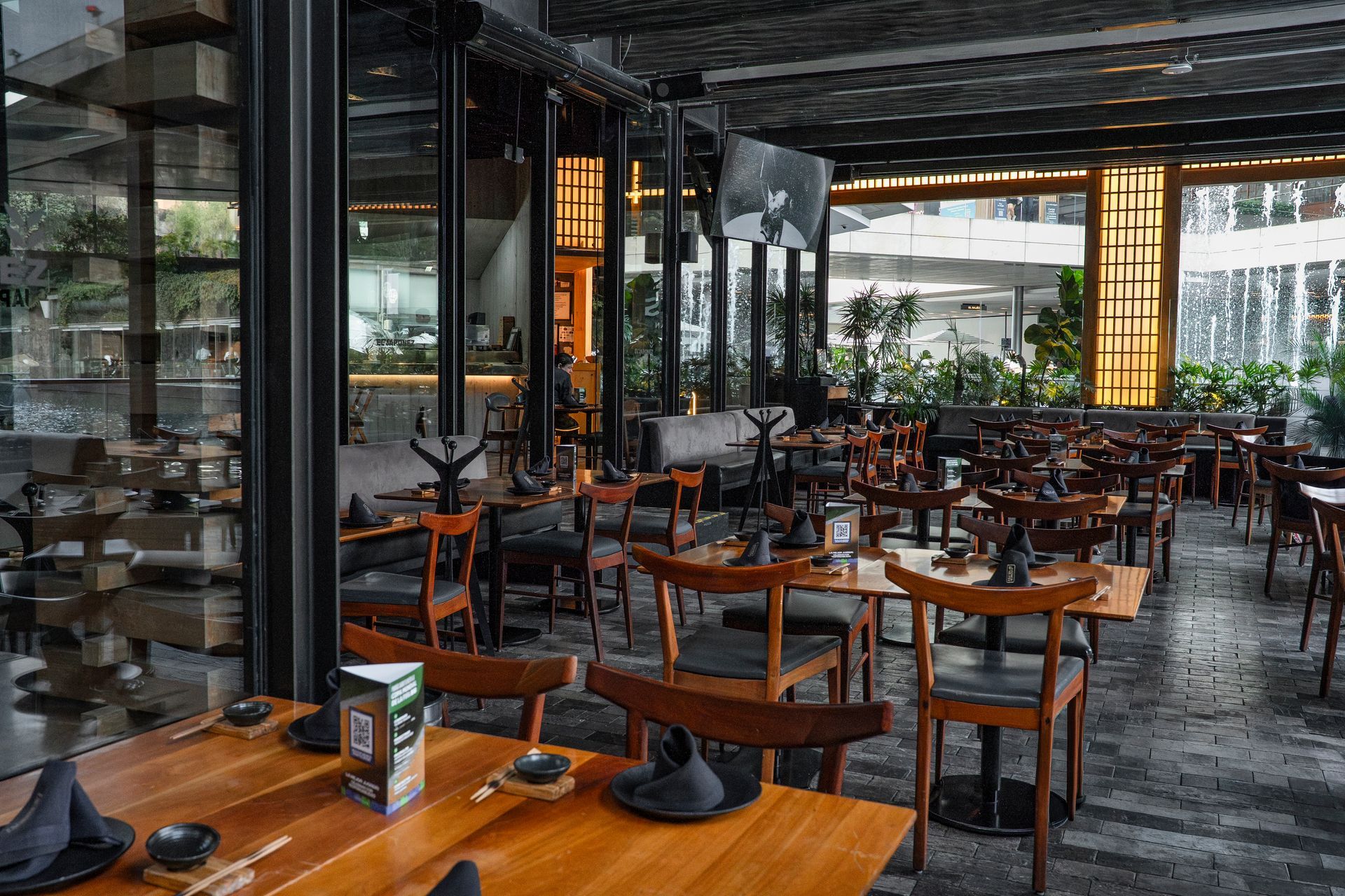 Restaurante moderno vacío con mesas de madera, sillas negras y paredes de cristal.