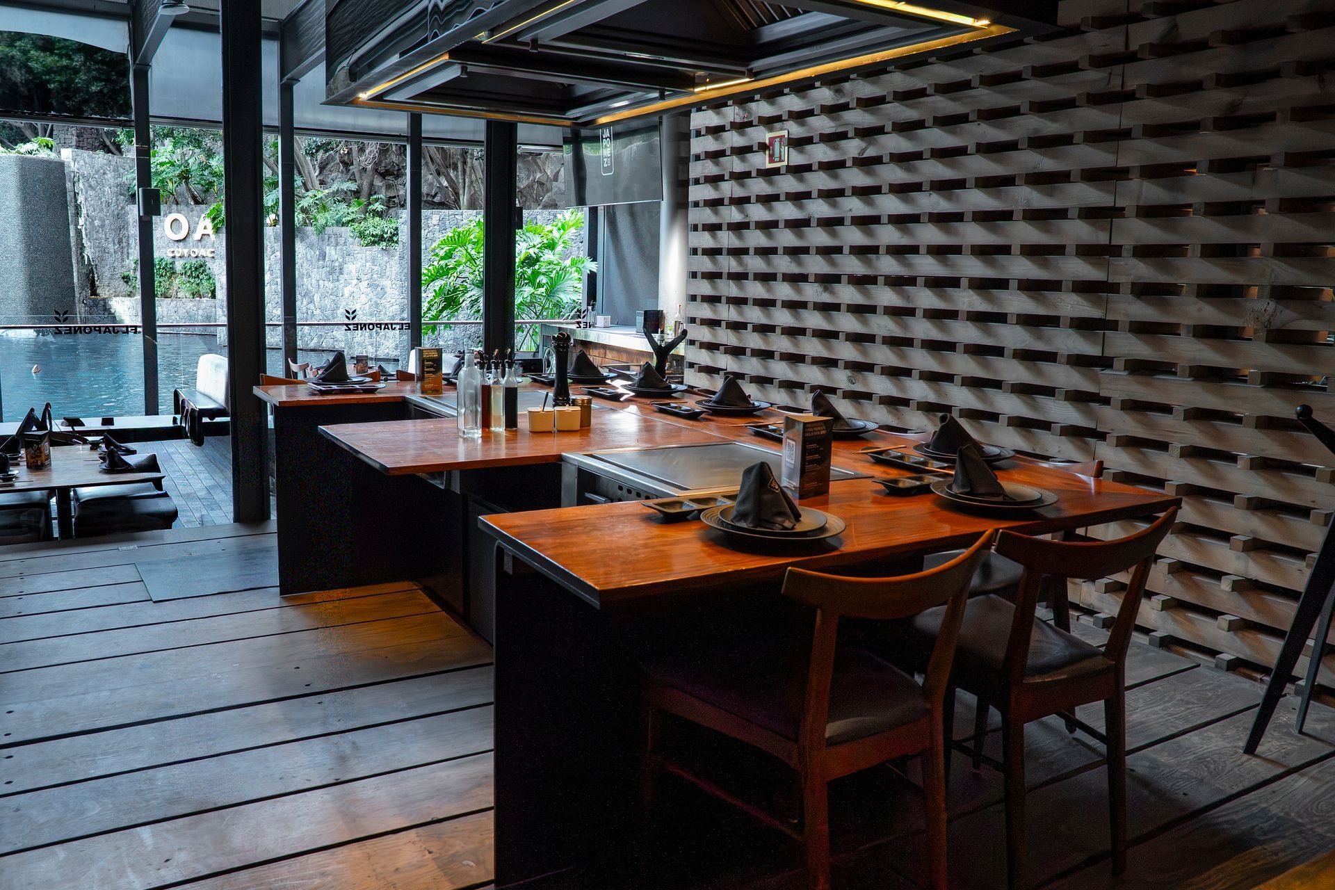 Interior de restaurante moderno con mesas y sillas de madera, y una pared estampada junto a grandes ventanales.