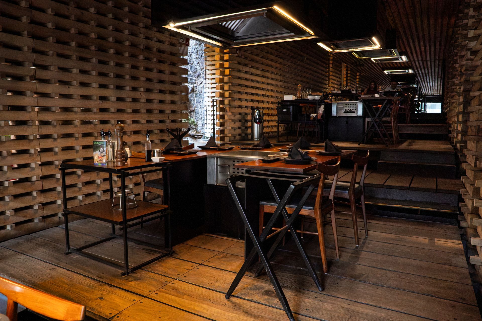 Interior de bar rústico con estantes de madera, mesas, taburetes y luces colgantes cálidas.
