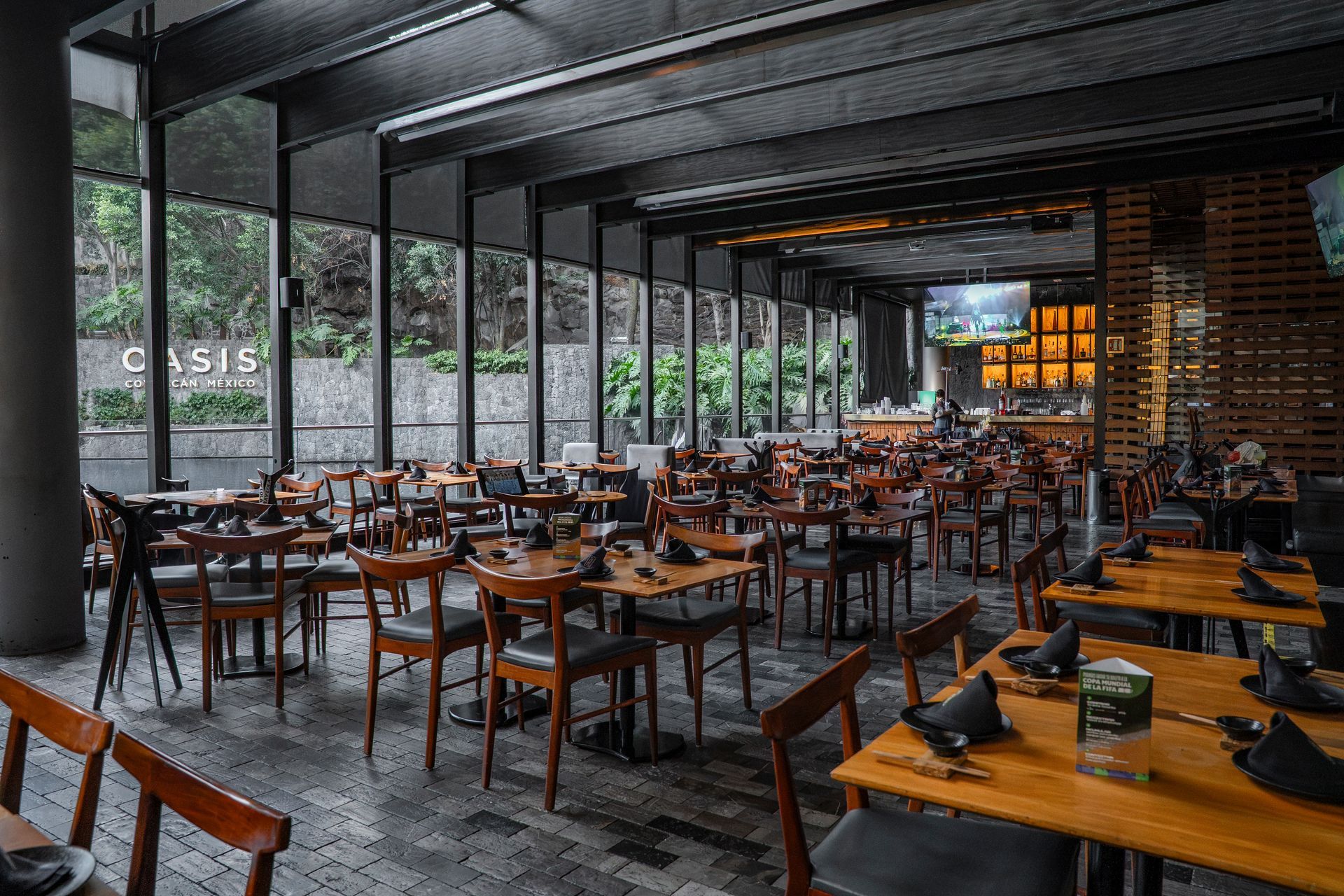 Interior de restaurante moderno con mesas y sillas de madera, y grandes ventanales con vistas a la vegetación.