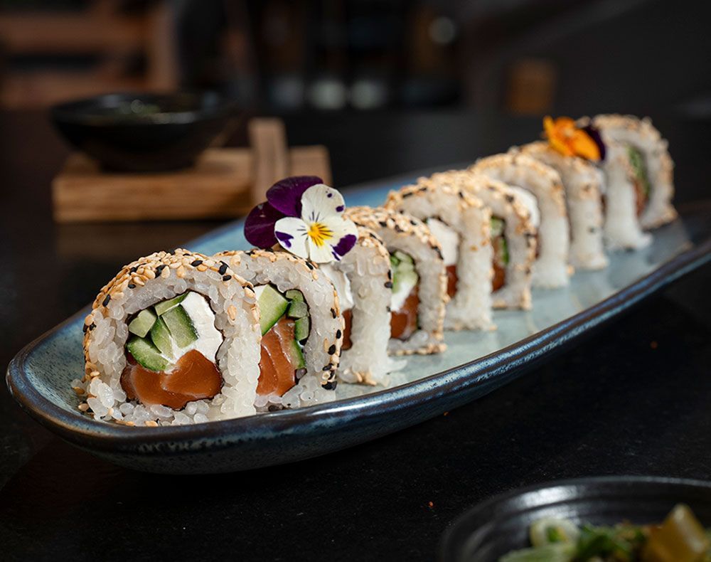 Rollos de sushi en un plato rectangular oscuro, adornados con flores comestibles, sobre una mesa de restaurante.