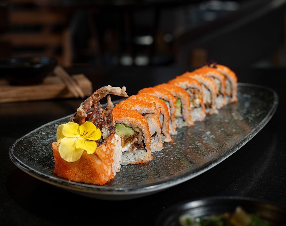 Rollo de sushi cubierto con salsa de naranja y cangrejo, servido en un plato negro con una flor amarilla como adorno.