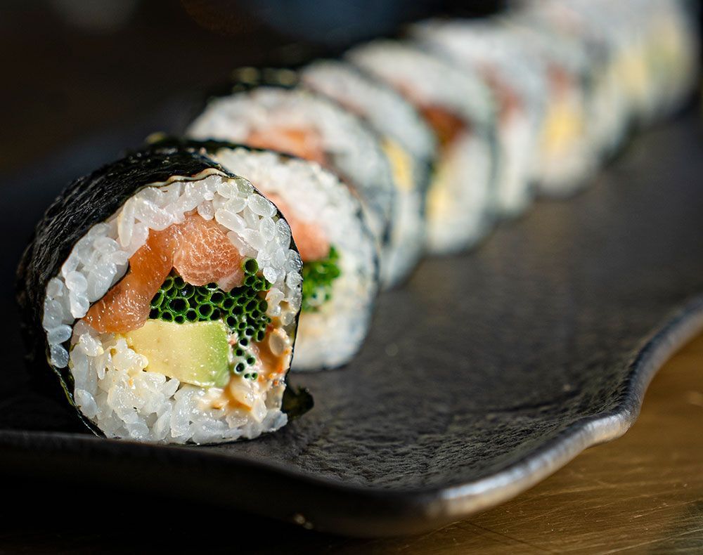 Primer plano de un rollo de sushi en un plato negro, donde se aprecian el salmón, el aguacate y las semillas de sésamo.