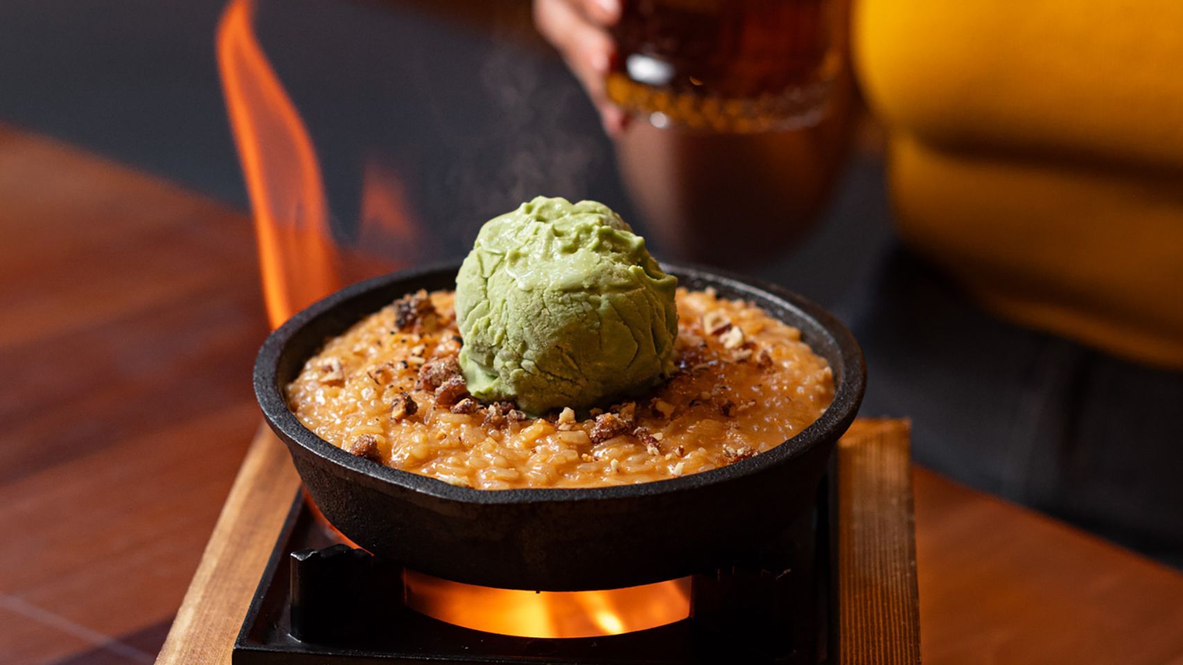 Postre en sartén cubierto con helado verde, flanqueado por llamas naranjas sobre una mesa de madera.