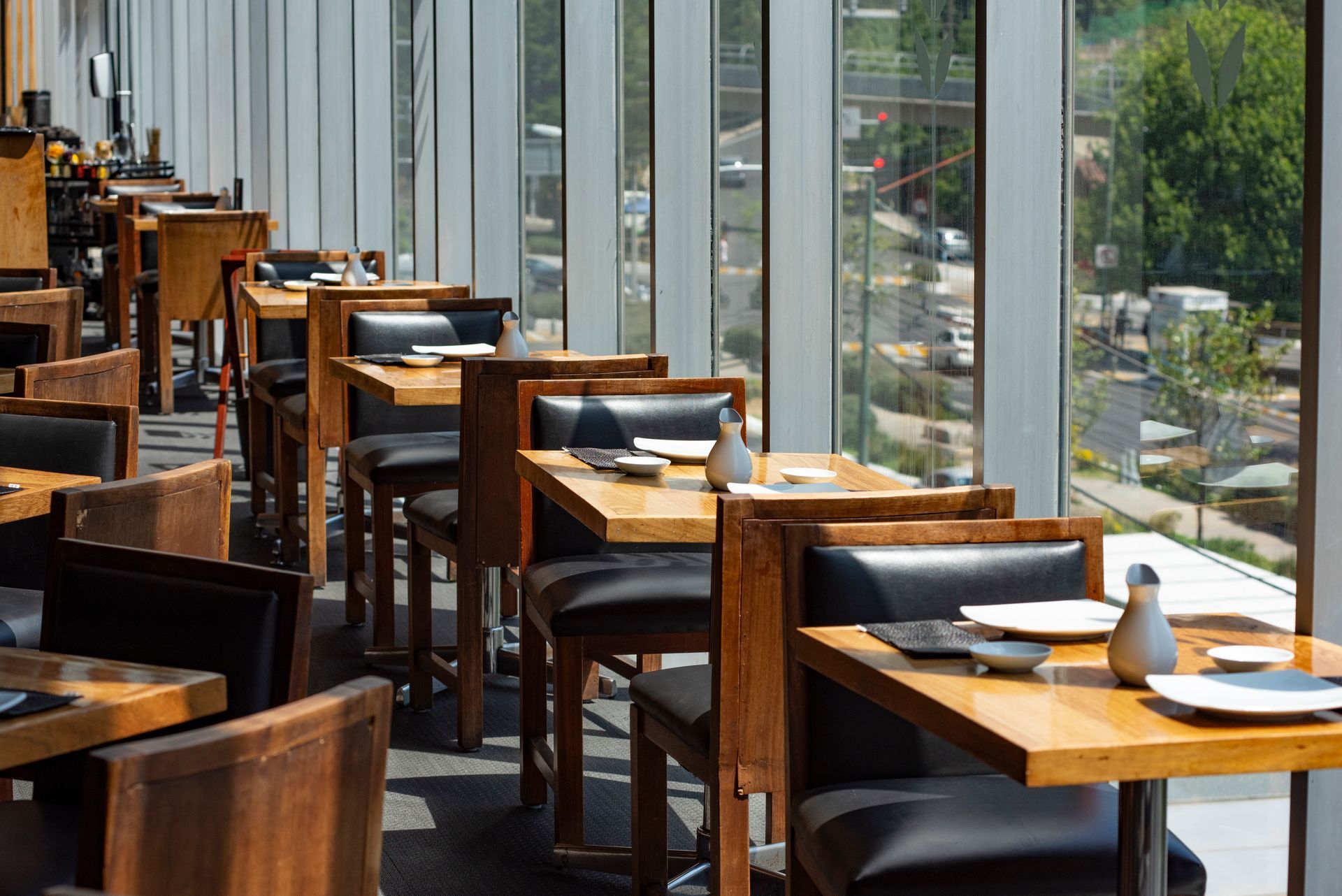 Comedor vacío con mesas de madera, sillas negras y grandes ventanales con vistas a la vegetación y la calle.