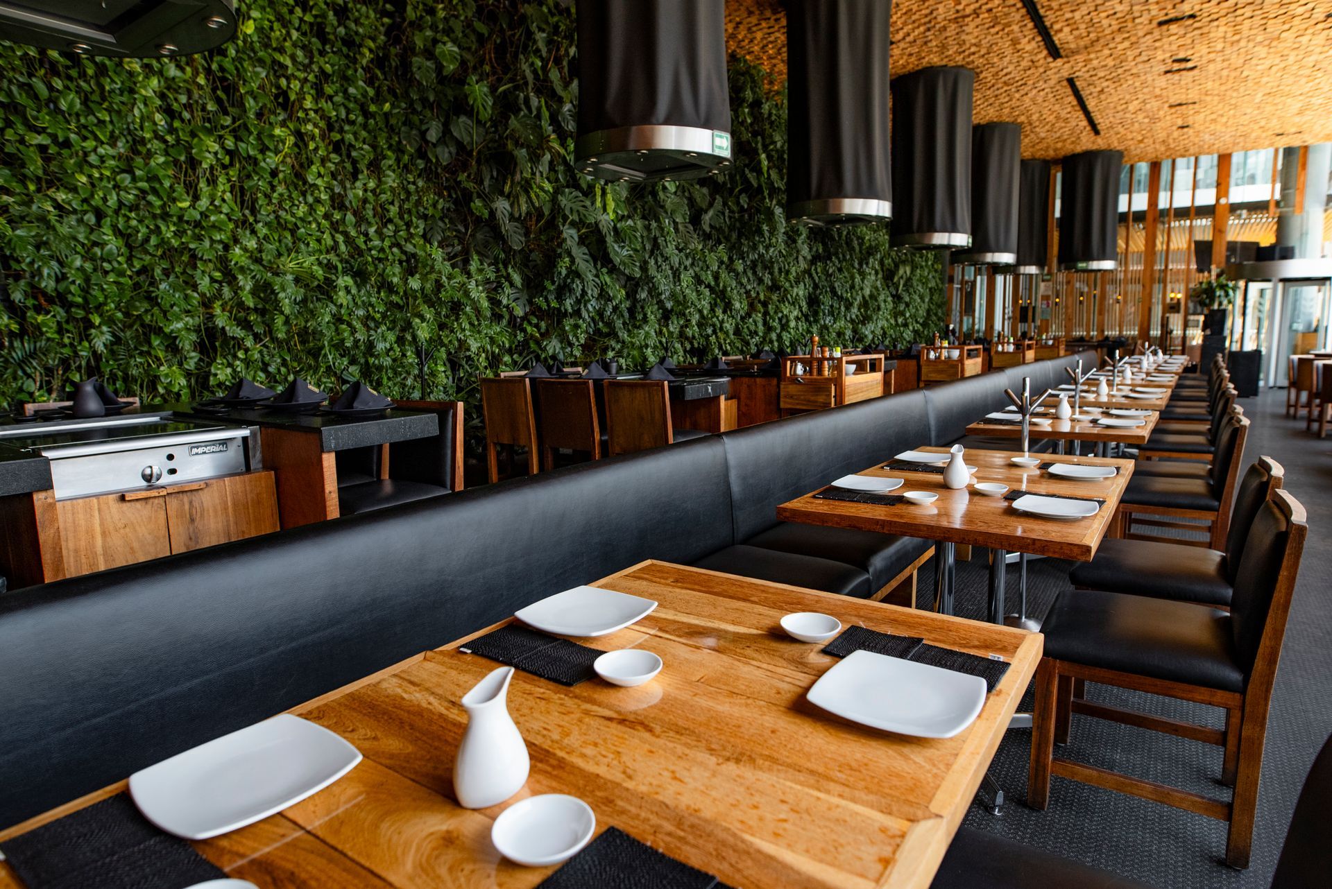 Amplio comedor de restaurante con mesas de madera, sillas negras y una pared vegetal verde.