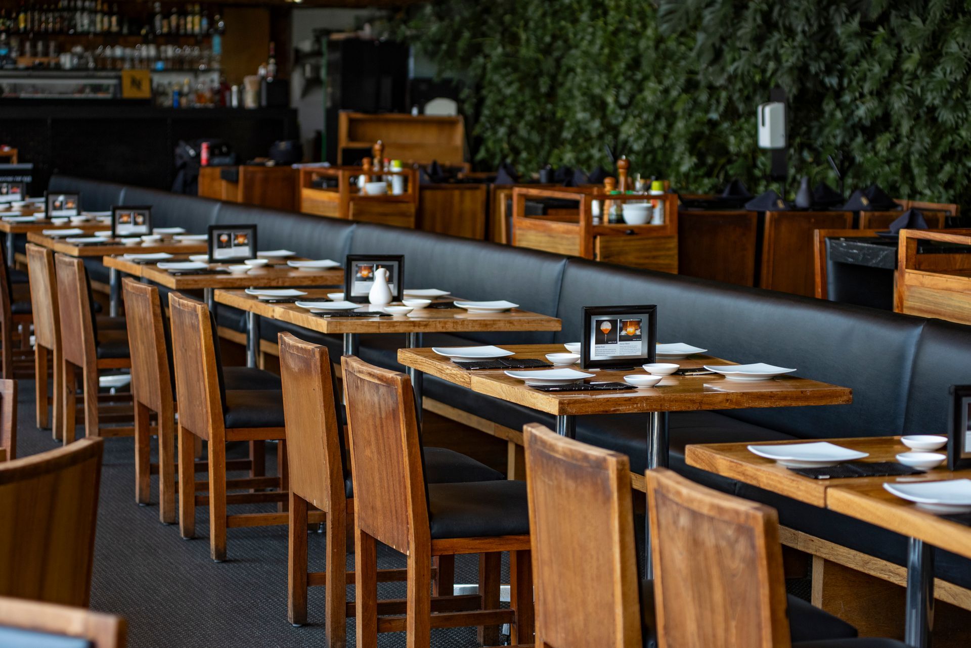Comedor vacío de un restaurante con mesas y sillas de madera, bancos grises y una pared cubierta de hojas verdes.