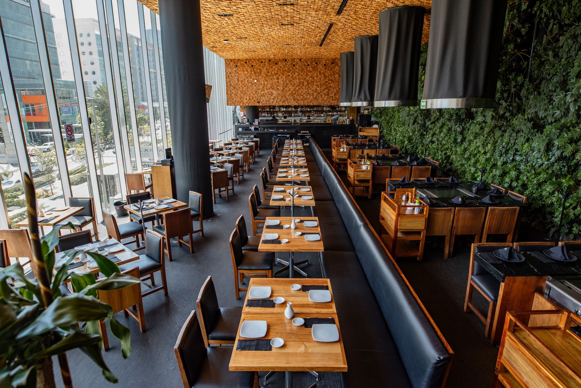 Interior de restaurante moderno con largas mesas de madera, asientos en la barra, vista a la ciudad y una pared verde.