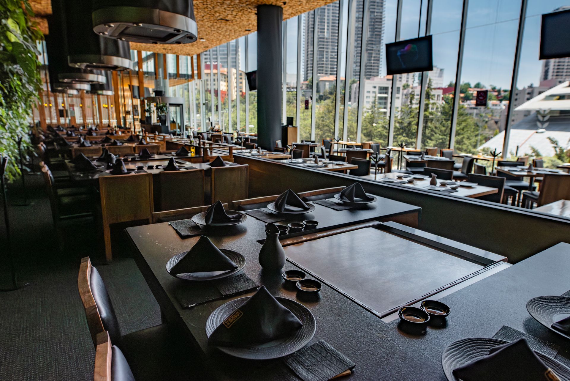 Comedor de restaurante moderno con mesas puestas, sillas negras y grandes ventanales con vistas a la vegetación.