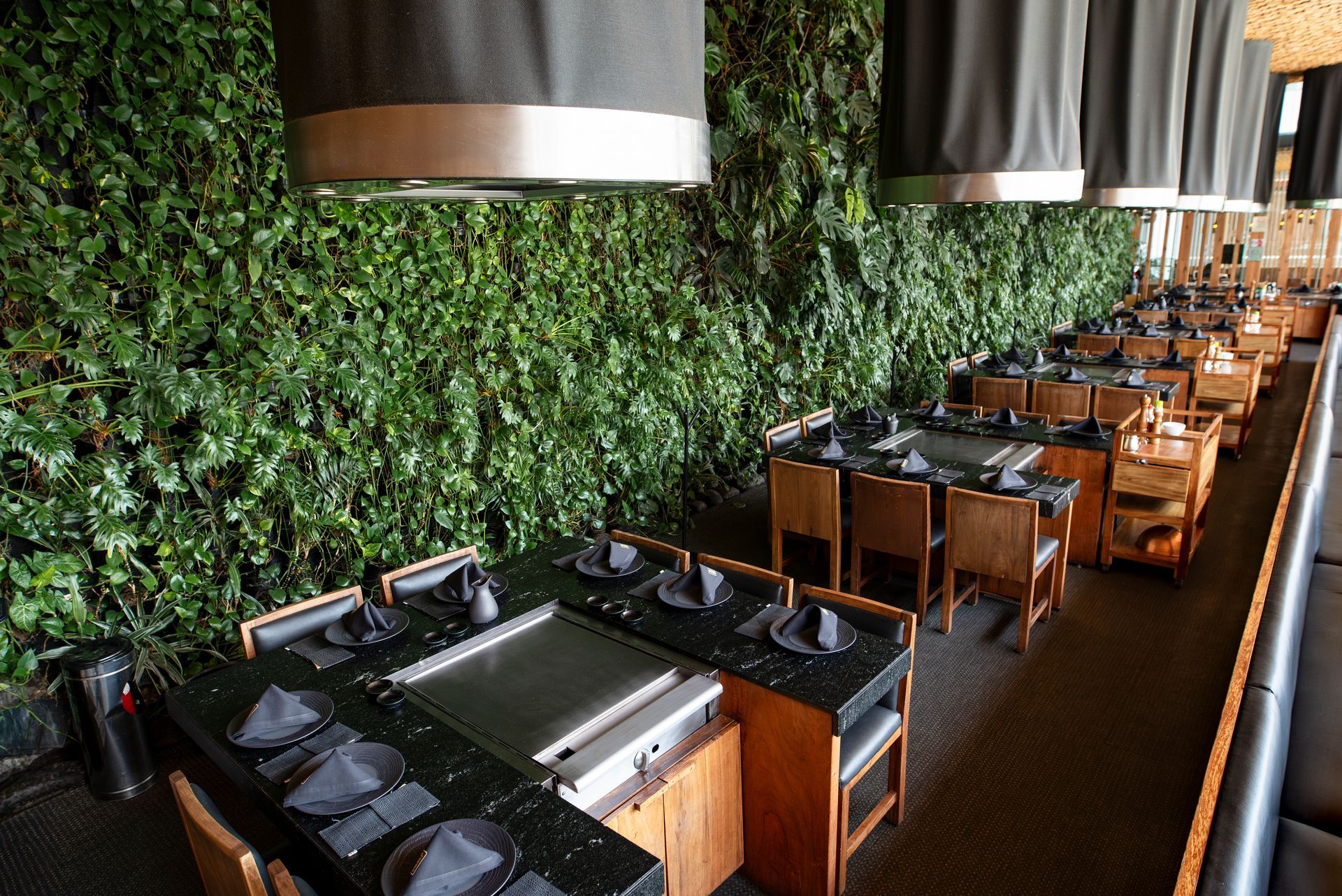Mesa de comedor larga en un restaurante elegante con una pared de plantas verdes y lámparas colgantes.