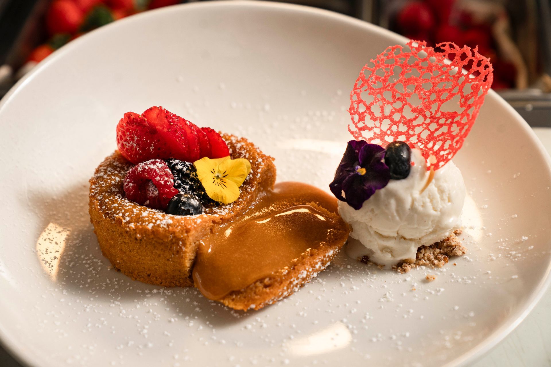 Postre gourmet en plato blanco con frutos rojos, salsa de caramelo, helado y guarnición de azúcar roja.