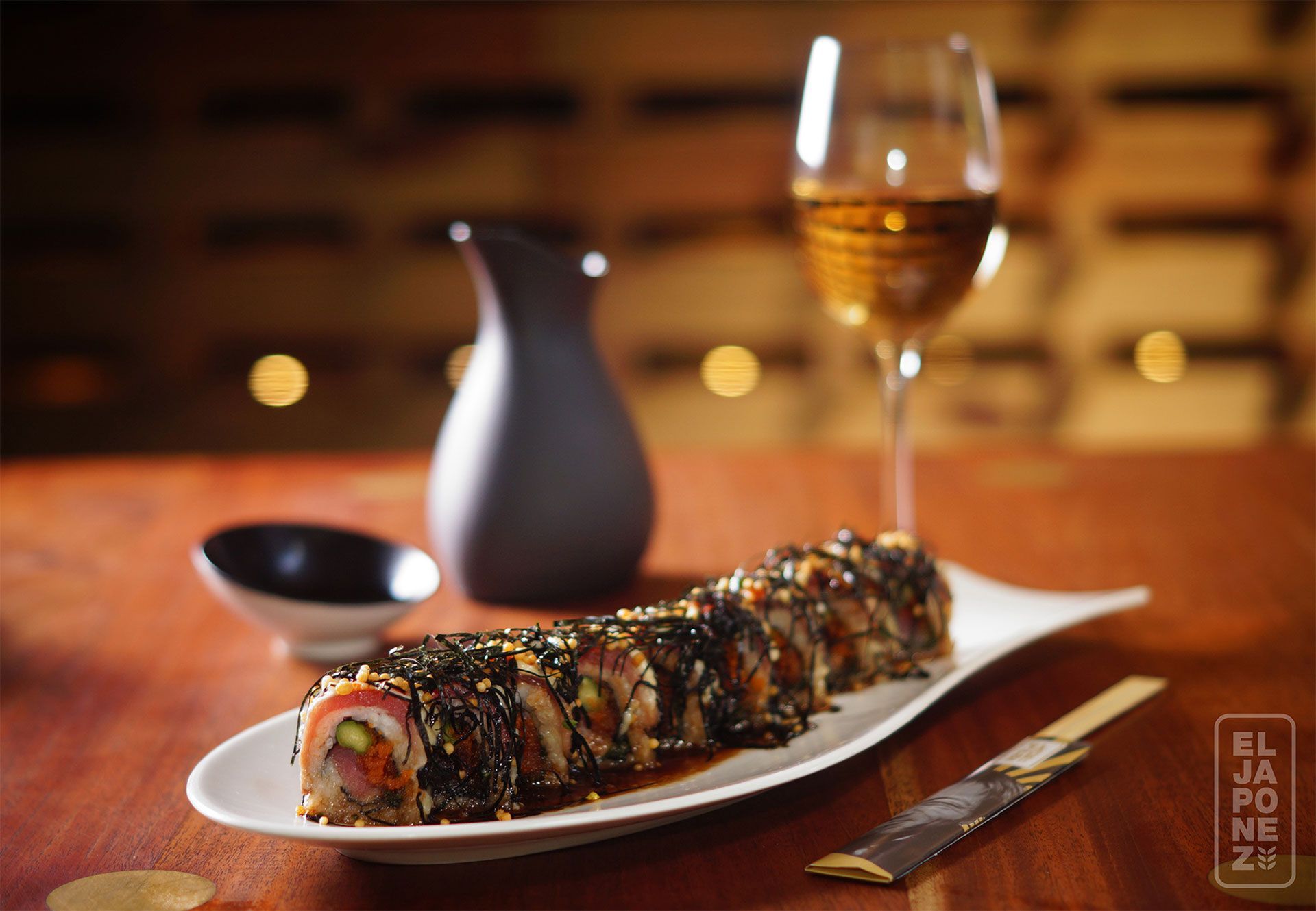 Un rollo de sushi en un plato blanco con una copa de vino y una jarrita en la mesa de un restaurante.