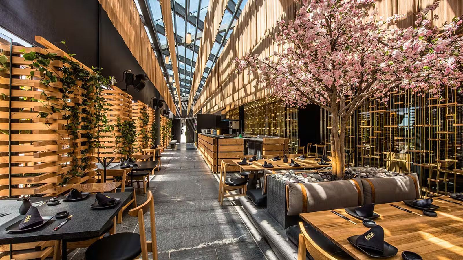 Interior de restaurante moderno con listones de madera, flores rosas y asientos a lo largo de un pasillo soleado.