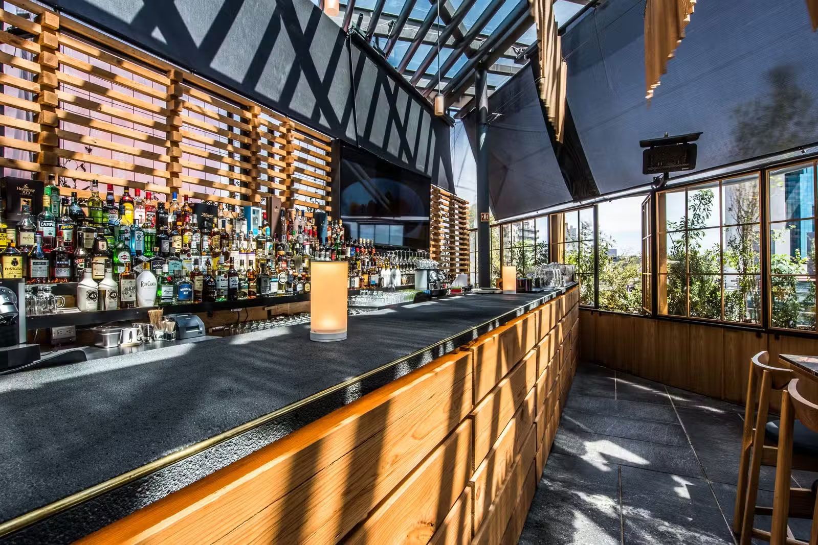 Interior de bar moderno con mostrador de madera, botellas y ventanas que dejan pasar la luz del sol.