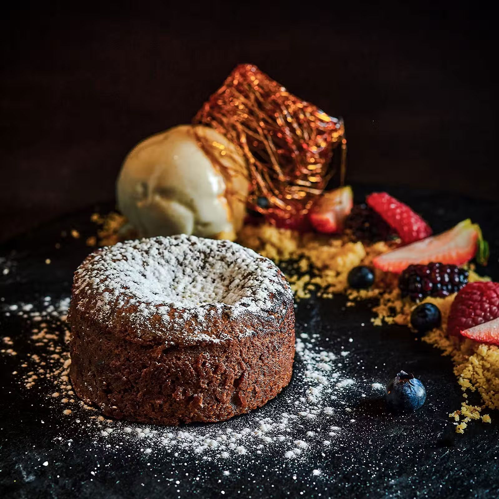 Postre de chocolate con azúcar glas, bola de helado, fresas y frutos rojos sobre una superficie oscura.