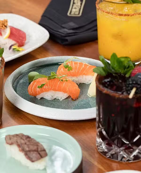 Sushi y postres en una mesa con dos bebidas coloridas y un cóctel oscuro.