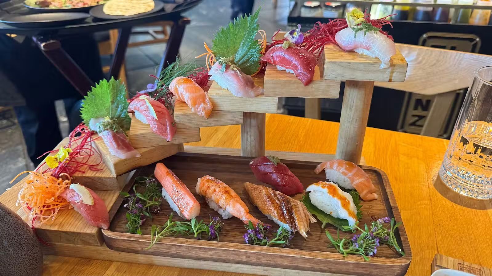 Plato de sushi con rollos variados y sashimi en una bandeja de madera, adornado con hierbas y flores rosas.