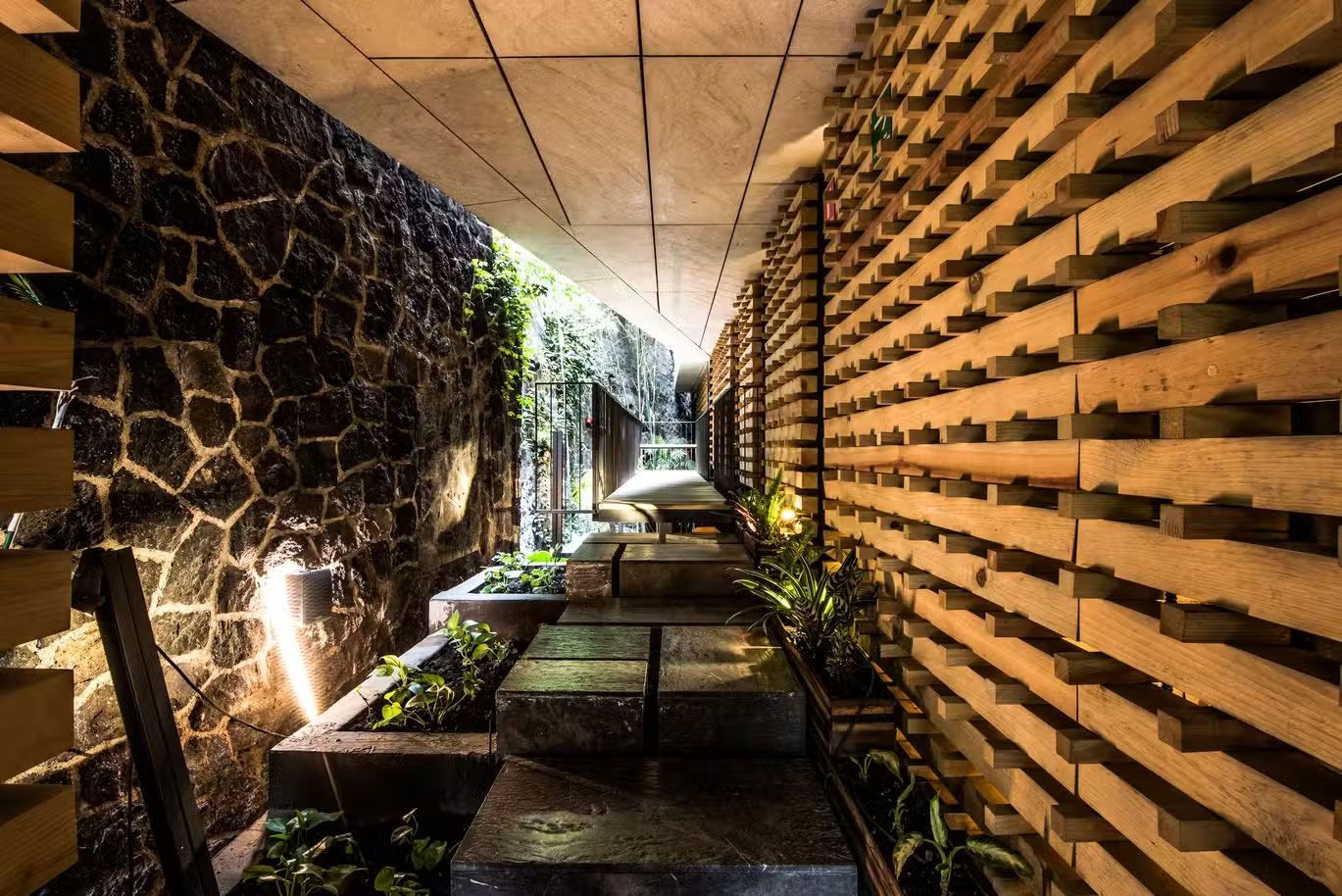 Un estrecho pasillo de piedra y madera con jardineras apiladas y cálidas luces de pared conduce a una exuberante vegetación.