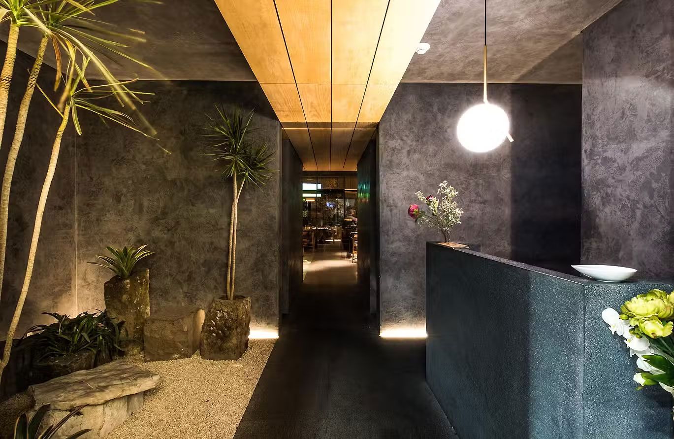 Pasillo moderno con paredes oscuras, iluminación cálida, mostrador de recepción y plantas en un interior minimalista.