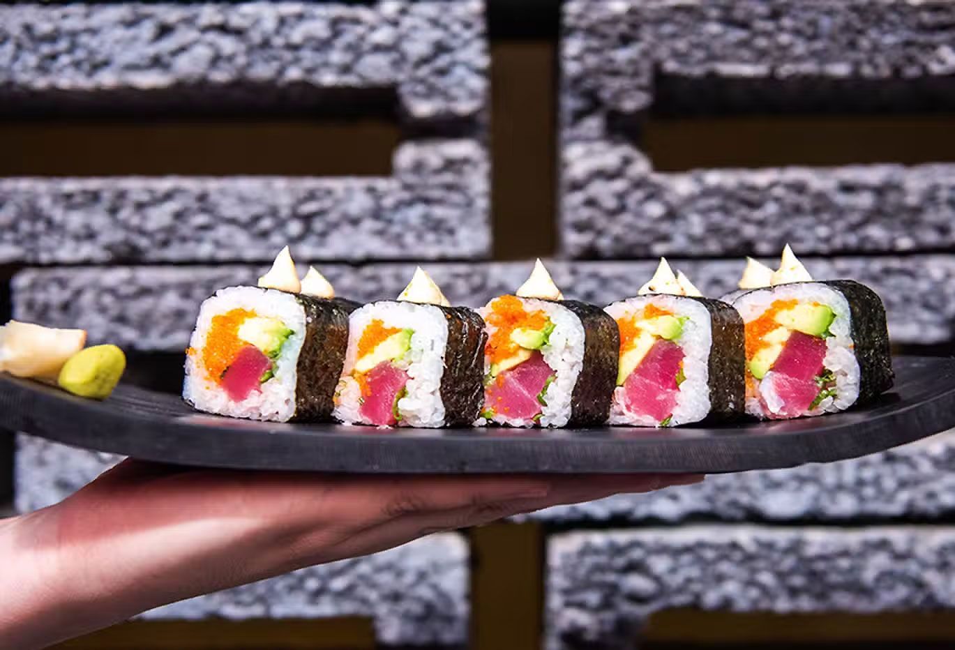 Rollos de sushi cubiertos con salsa blanca en un plato negro sostenido por una mano contra un fondo de pared de ladrillos.