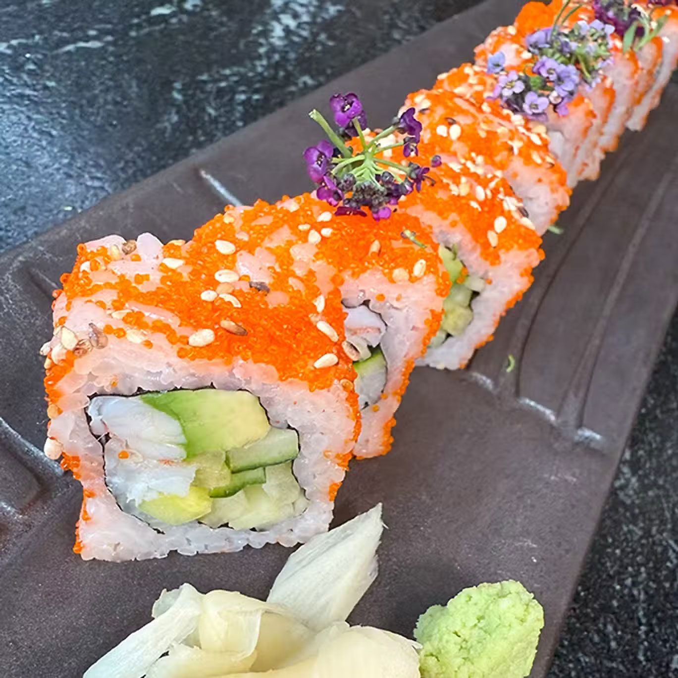 Rollos de sushi cubiertos con huevas de naranja y hierbas en un plato oscuro, con wasabi y jengibre encurtido.