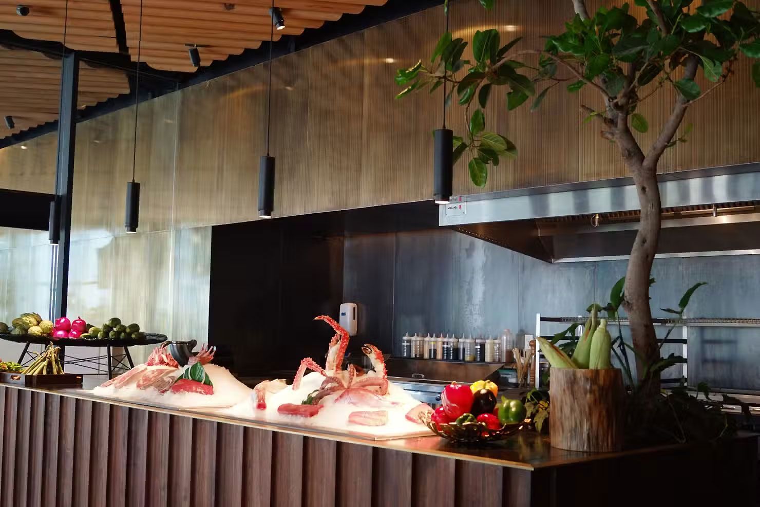 Barra de buffet con bandejas de mariscos, flores y un árbol en el interior de un restaurante moderno.