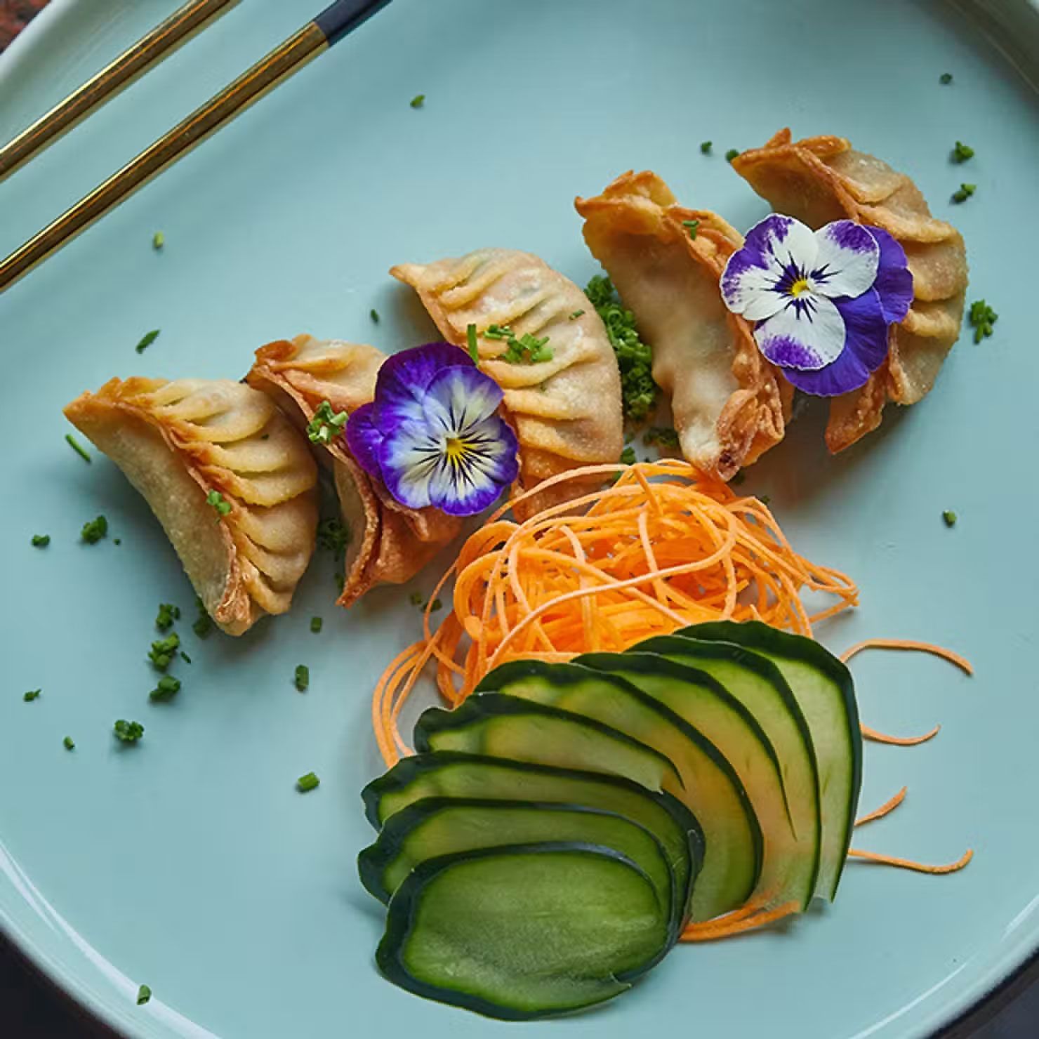 Empanadillas emplatadas con flores moradas, rodajas de pepino y guarnición de zanahoria en un plato azul claro.