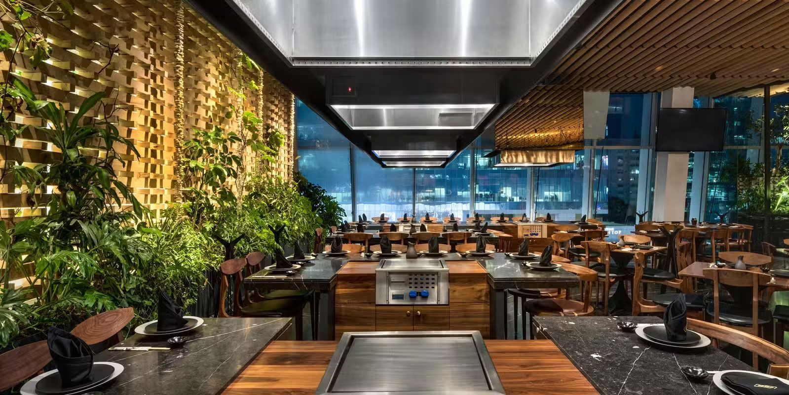 Interior de restaurante moderno con iluminación cálida, mesas de madera y decoración de pared de exuberante vegetación.