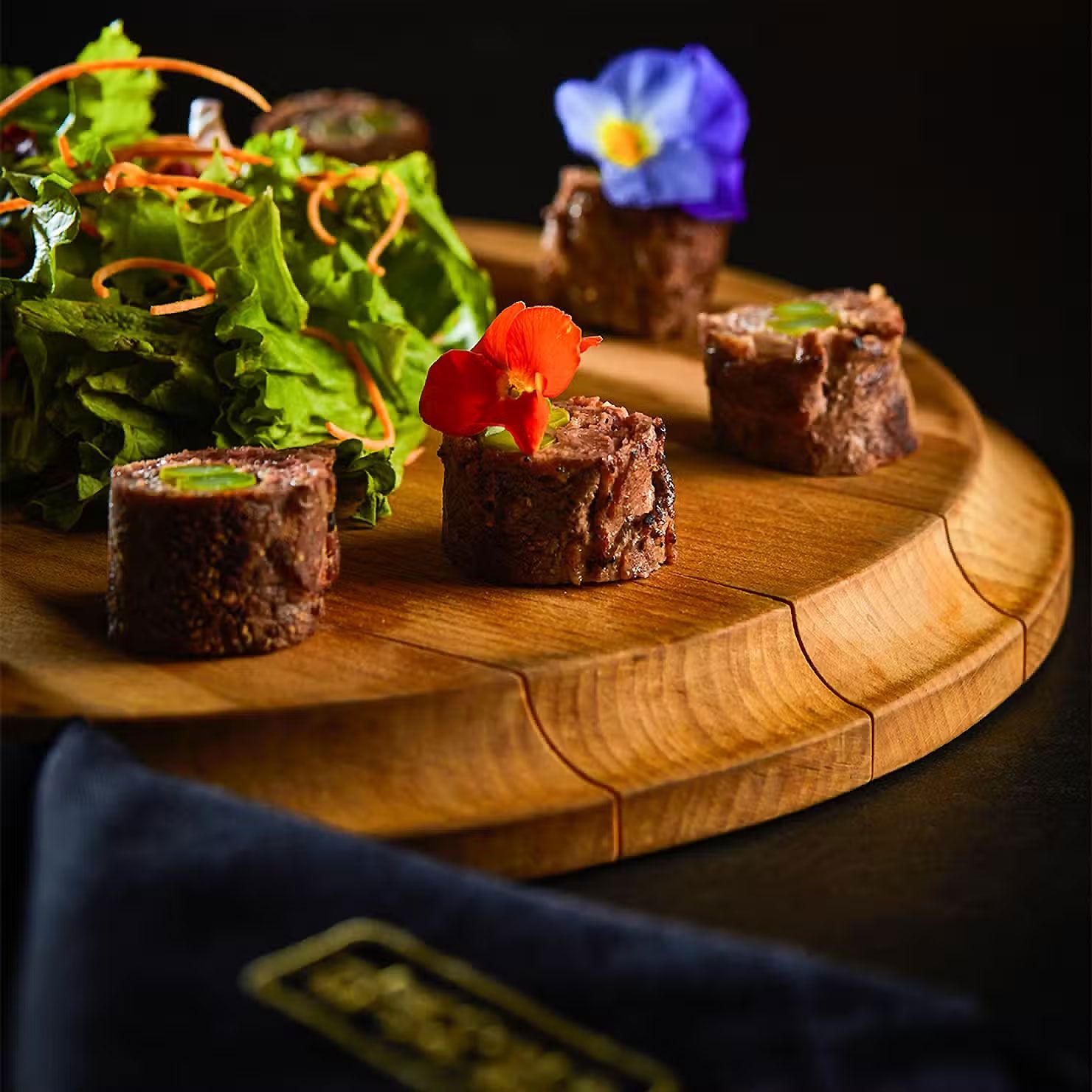 Bocaditos gourmet de carne servidos en una tabla de madera con verduras y flores comestibles.