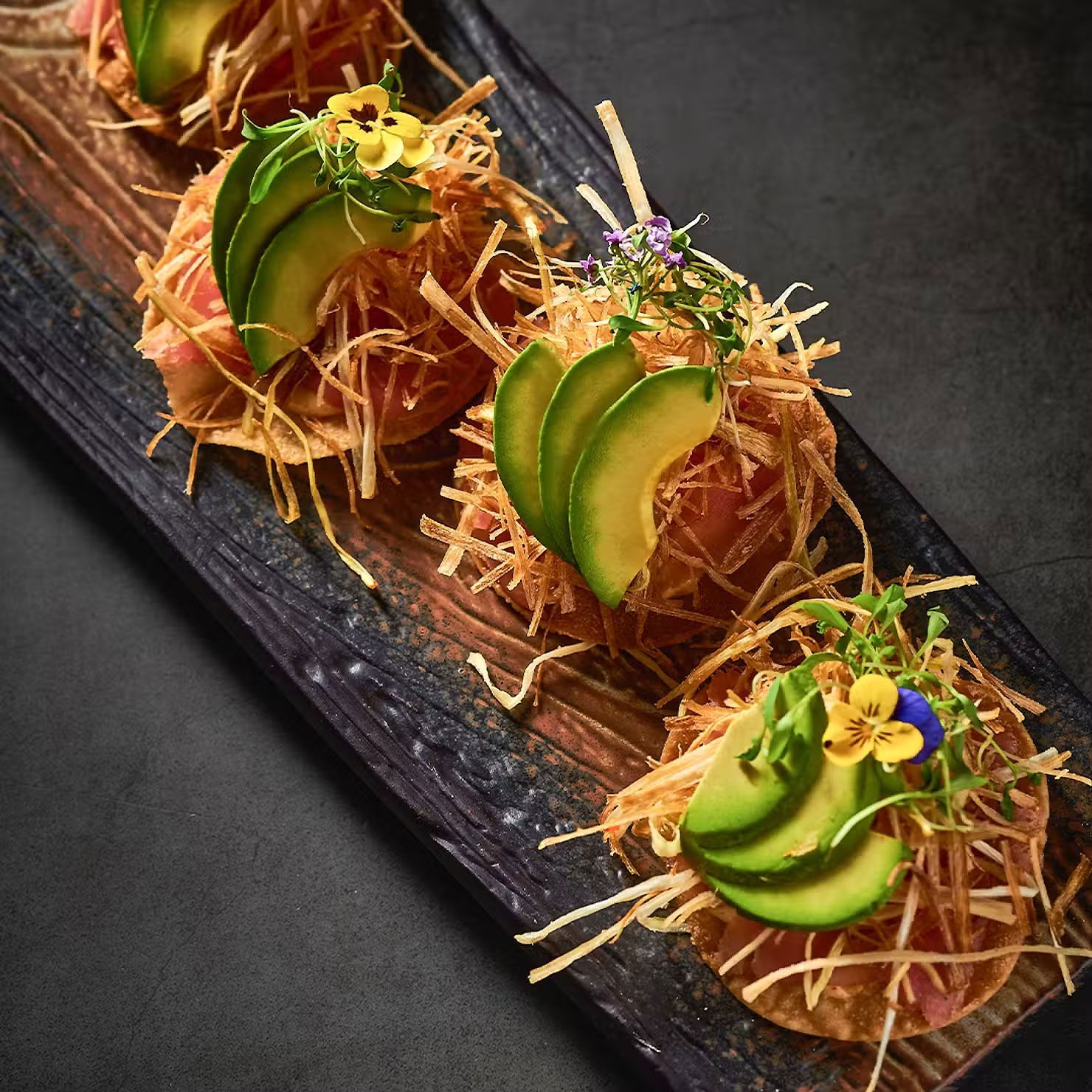 Tres postres gourmet en una bandeja de pizarra, decorados con rodajas de kiwi y flores comestibles.