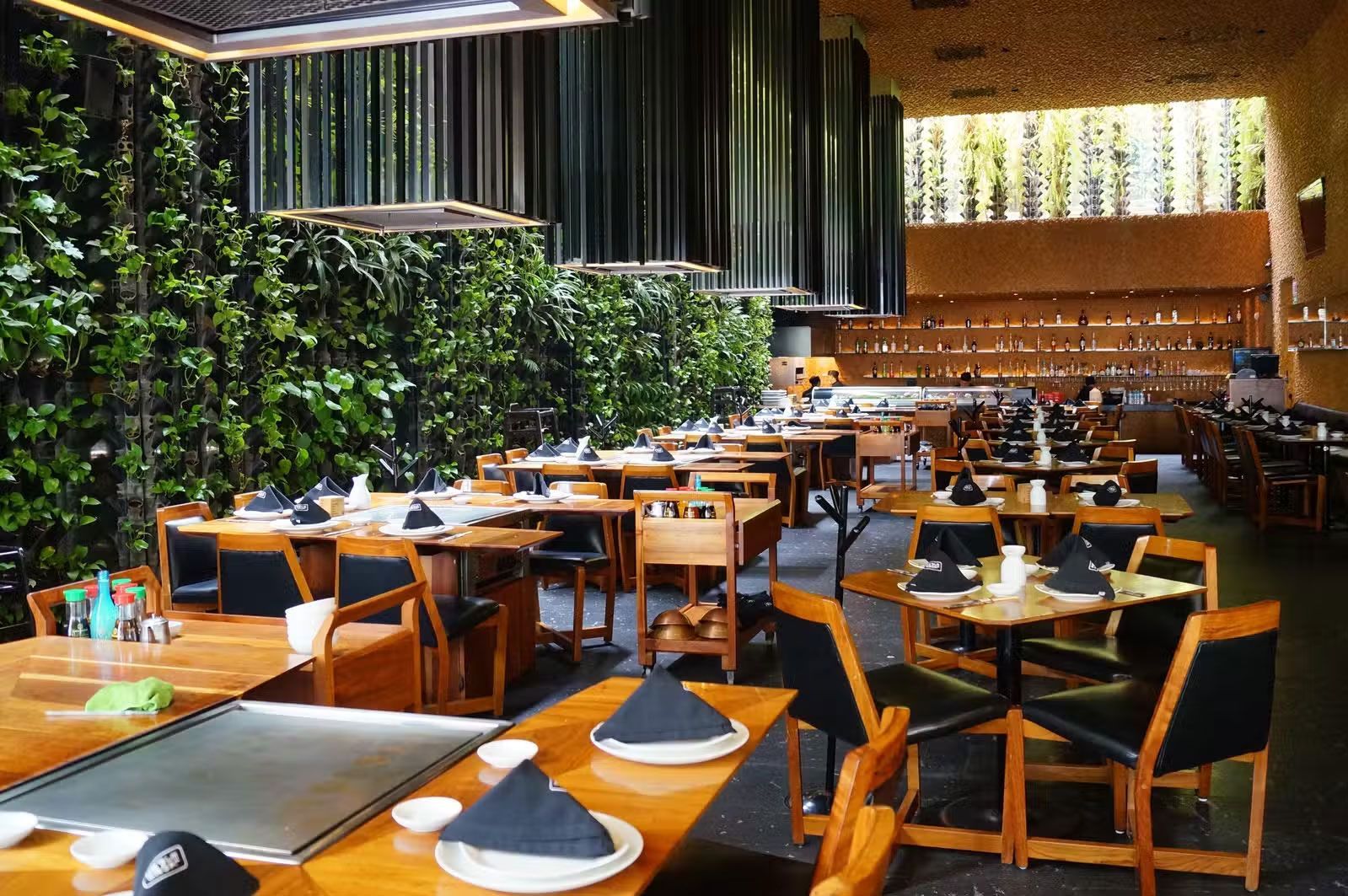 Comedor vacío de un restaurante de lujo con mesas de madera, sillas negras y una pared vegetal verde.