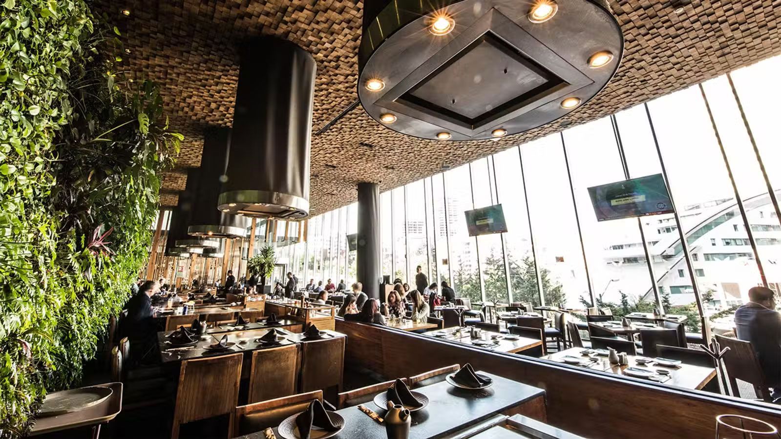 Interior de restaurante moderno con vegetación, mesas de madera y grandes ventanales con vistas a la calle.