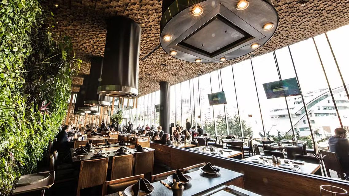 Interior de restaurante moderno con mesas de madera, plantas colgantes y grandes ventanales con vistas a la ciudad.