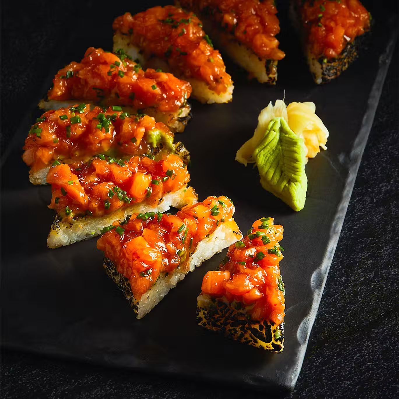 Rollos de sushi cubiertos con salmón picante en un plato de pizarra negra, adornados con wasabi.