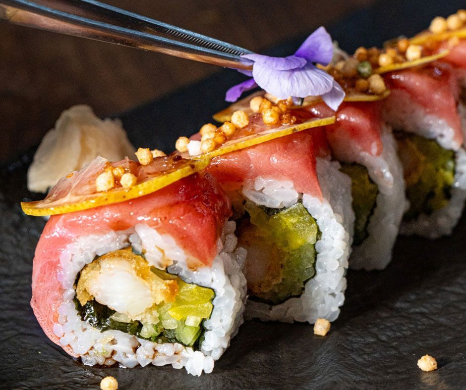 Rollos de sushi cubiertos con salmón, caviar y flores comestibles en un plato oscuro.