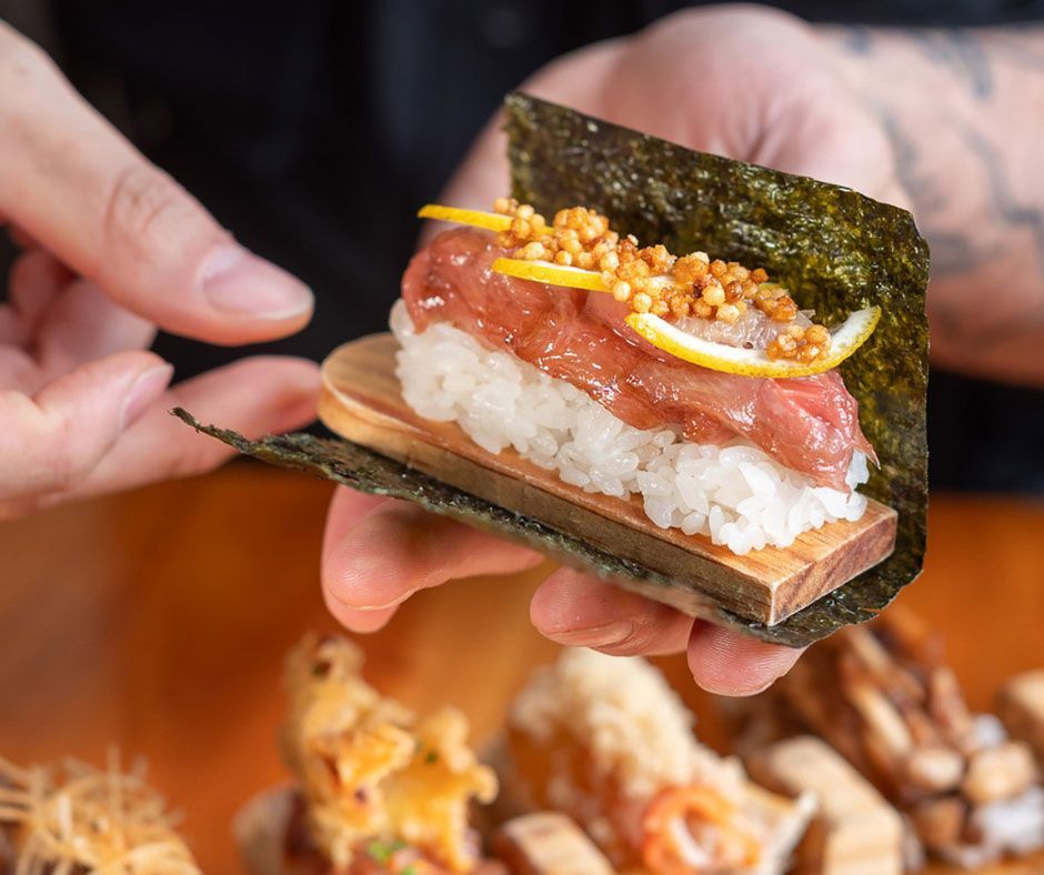 Manos preparando un rollo de sushi con alga nori, arroz, salmón y una cobertura crujiente.