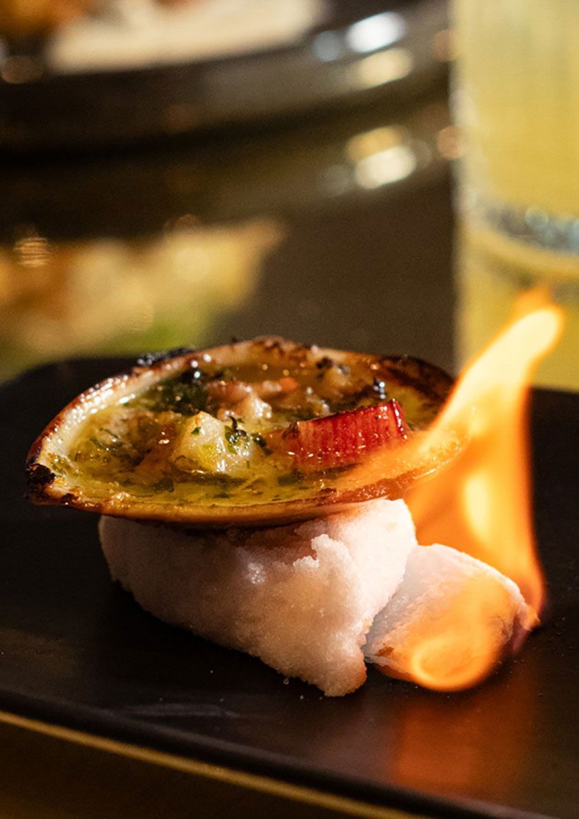Plato horneado flameado sobre un plato negro, cubierto con hierbas y una rodaja de limón.