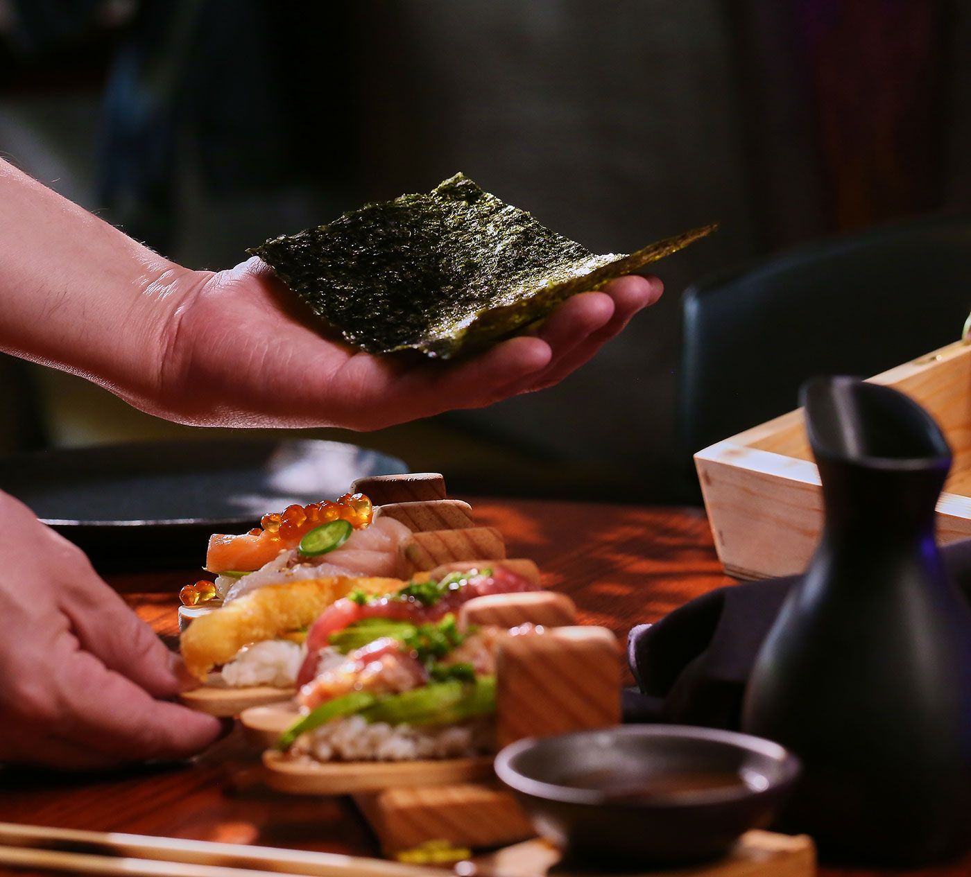 Colocación manual de una lámina de alga marina sobre rollos de sushi en una bandeja de madera.